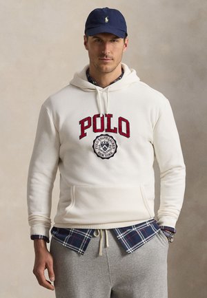 Homme portant un sweat à capuche blanc Polo Ralph Lauren, chemise à carreaux bleue, pantalon de survêtement gris, casquette bleu marine et montre-bracelet, debout devant un fond neutre.