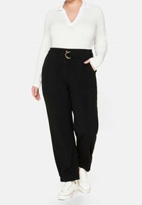 Žena v černých kalhotách s vysokým pasem a páskem, bílém dlouhém rukávu s límečkem a bílých teniskách, stojící s jednou rukou v kapse.