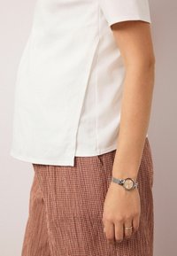 Weißes Kurzarmhemd mit Seitenschlitzen, kombiniert mit braunen, karierten Hosen. Silberne Mesh-Armbanduhr mit rundem Zifferblatt und hellem Ziffernblatt.