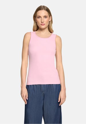 Femme portant un haut sans manches rose clair associé à un pantalon ample bleu foncé, debout devant un fond clair uni.