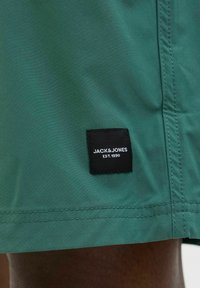 Žalios šortai pagaminti iš lygaus audinio; turi siuvinėtą kraštą ir juodą etiketę su užrašu "JACK & JONES EST. 1990".