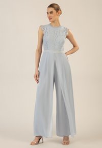 Himmelblauer Jumpsuit mit Spitzentop und kurzen Ärmeln, plissierter, weit geschnittener Design, weicher Stoff und taillierter Schnitt. Silberne Sandalen runden den Look ab.