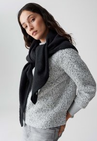 Schwarzer, übergroßer Schal, der um einen hellgrauen, strukturierten Pullover drapiert ist, ergänzt durch einen gerippten Ärmelsaum, vor einem neutralen Hintergrund.