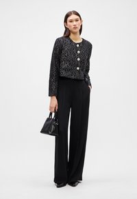 Giacca corta nera con motivo floreale argentato, dotata di tre grandi bottoni. Abbinata a pantaloni neri a vita alta e una piccola borsa.