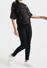 Persona vestida con una camiseta de manga corta con patrón de manchas negras, jeans ajustados negros y zapatillas blancas, de pie con los brazos ligeramente extendidos.