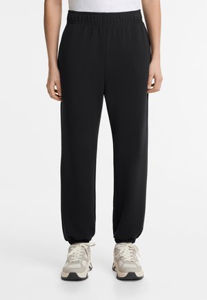 Black, elastic-waist jogger pants with tapered cuffs and a smooth texture, paired with light-colored sneakers featuring multiple layers.