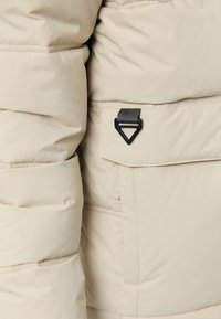 Beige pufferjack detail met een geometrisch quilted textuur, met een zijzak met een zwart driekhoekig hardwareaccent en gestikte naden.
