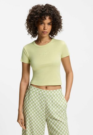 Mujer con cabello rizado que lleva una camiseta corta verde claro con el texto "Hugo" y pantalones verdes y blancos con estampado, parada con una mano en el bolsillo.