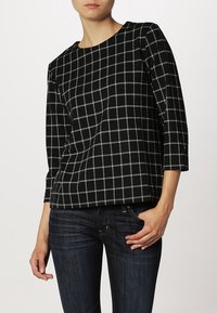 Žena oblečená v černém svetru s bílým mřížkovaným vzorem a tříčtvrtečními rukávy, v kombinaci s tmavě modrými džínami, s rukou v kapse.