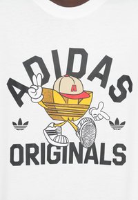 Balti marškinėliai su geltonos spalvos animacinio personažo grafiniu dizainu su kepuraite, šalia stambiu juodu šriftu užrašyta „ADIDAS ORIGINALS“.