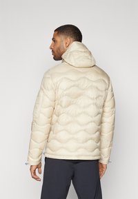 Padded jacket in cream color with a hood, featuring a wave-patterned texture and blue accents on the cuffs. Smooth fabric finish.