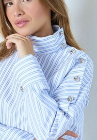 Blå och vit randig skjorta med hög krage, detaljerade med silverknappar på axel och manschett, tillverkad av ett slätt tyg med skräddarsydd design.