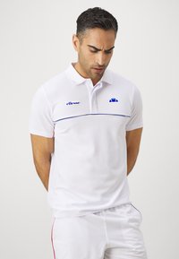 Hvit kortermet polo-skjorte med strukturert stoff, blå logo og horisontal blå stripe over brystet. Brukt med hvite shorts.