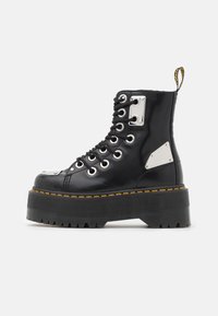 Botas de tobillo de charol negro con herrajes metálicos, diseño de cordones, suela gruesa y acento de costura amarilla.