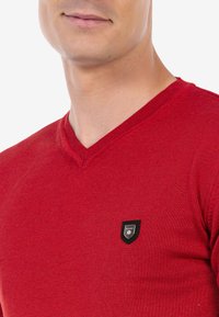 Roter V-Ausschnitt-Pullover aus Strickstoff, mit einem kleinen schwarzen Shield-Logo auf der linken Brust. Die Textur wirkt glatt und eng anliegend.