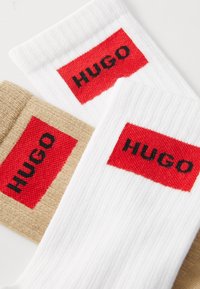 Biele a béžové ponožky s rebrovanou textúrou, pričom každé z nich má červený obdĺžnikový záhyb s nápisom "HUGO" tučnými čiernymi písmenami.