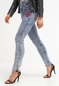 Chude dżinsy w jasnoniebieskim odcieniu z kwiatowym haftem w kolorze różowym i fioletowym. Posiadają rozcięcia na końcach i przetarcia. Stylizowane z czarnymi szpilkami.
