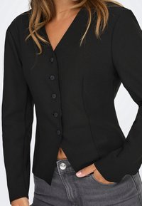 Femme portant un blazer noir cintré boutonné à manches longues et un jean gris, une main dans la poche, cadrée à la taille.