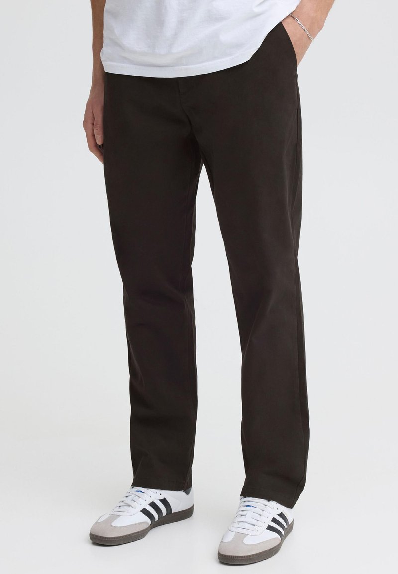 Homme portant un pantalon droit noir, des baskets blanches avec des bandes noires et un t-shirt blanc, debout avec une main dans la poche.