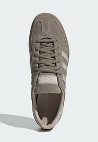 Γκρι σουέντ αθλητικό παπούτσι με τρεις ανοιχτό γκρι ρίγες, υφασμένα κορδόνια και λαστιχένια σόλα· διαθέτει ετικέτα στη γλώσσα με λεπτομέρειες εμπορικού σήματος.