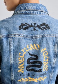 Džinsinė striukė su juodu gėlių raštu ant apykaklės ir dideliu juodu gyvatiškos grafika, apsupta geltonų tekstų gale.