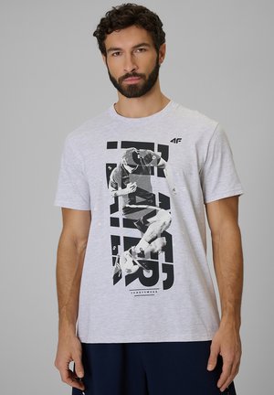 Mężczyzna z ciemnymi kręconymi włosami i brodą, ubrany w jasnoszary t-shirt z czarno-białą grafiką piłkarza i granatowe szorty na szarym tle.