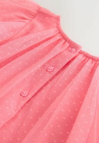 Tissu rose transparent à petits pois, encolure froncée et trois boutons roses le long du centre avant.