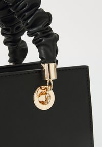 Sac à main noir en cuir lisse. Il présente une anse froncée et une quincaillerie dorée avec une attache circulaire.