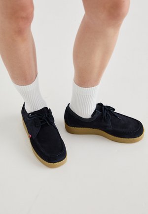 Marineblaue Wildlederschuhe mit runder Zehenpartie, Schnürdesign und cremefarbener Gummisohle. Kombiniert mit weißen gerippten Socken.