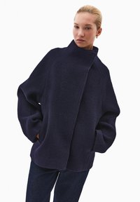 Granatowy, oversize'owy płaszcz wykonany z teksturowanej tkaniny, z wysokim kołnierzem i dużymi kieszeniami. Detale szwów podkreślają drapowaną sylwetkę.
