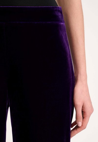 Paarse fluwelen broek met een zachte textuur, voorzien van een brede tailleband en een gladde afwerking, waarbij een hand aan de zijkant steunt.