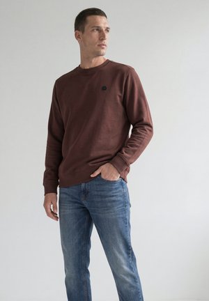Mand iført brun langærmet sweatshirt og blå jeans, stående med den ene hånd i lommen, kigger til siden mod en ensfarvet baggrund.