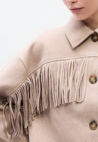 Beežist villast jope, millel on krae, ees nupud ning rinnal ja õlgadel pikad narmad, lisades tekstuuri ja detaile.