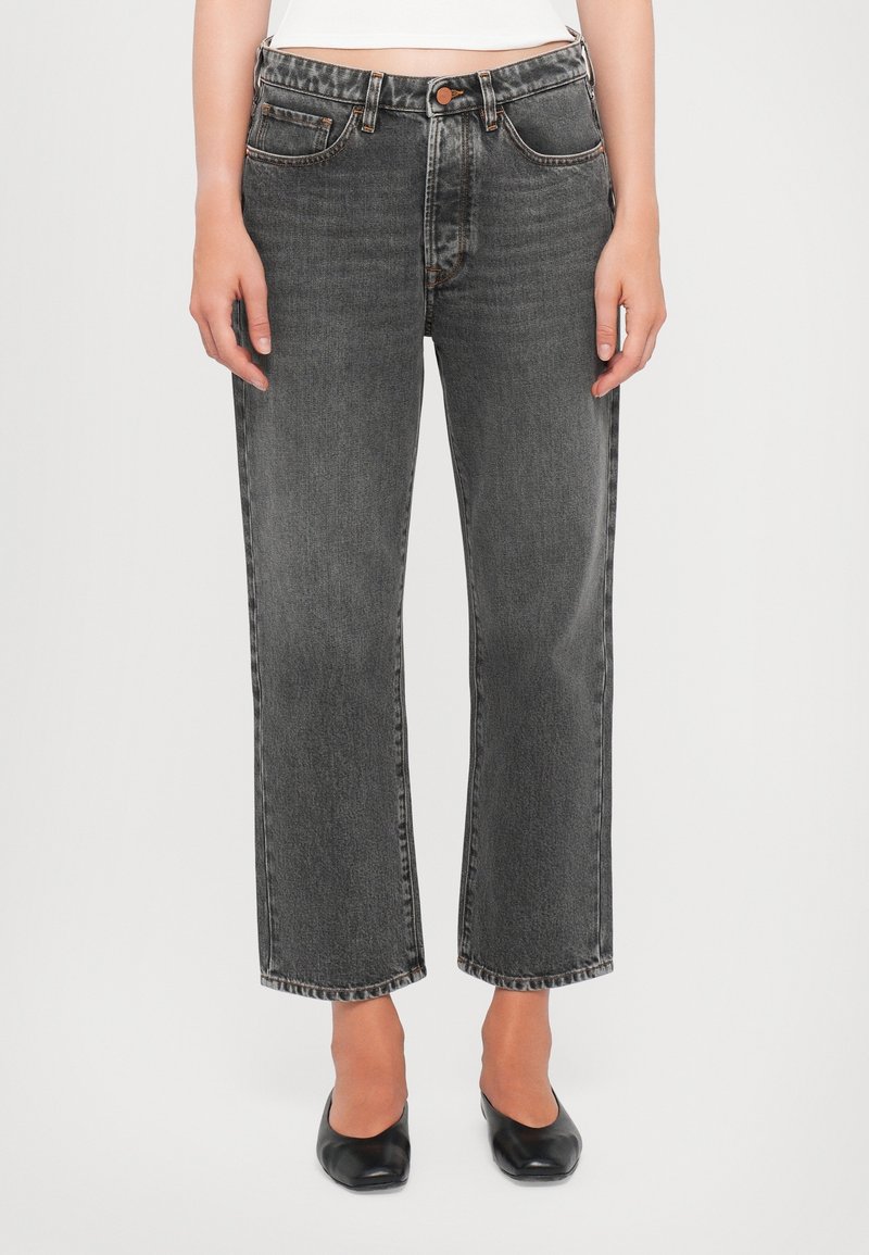 Femme portant un jean noir délavé à jambes droites et des chaussures noires à bout carré, debout devant un fond blanc.