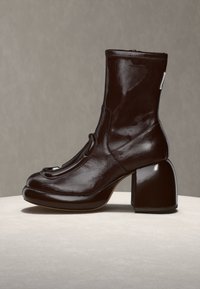 Bottine en cuir brun foncé brillant avec bout carré arrondi et talon bloc large sur une surface et un fond neutres.