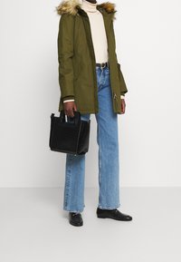 Person iført en olivengrøn parka med pelskrave, cremet turtleneck, lyseblå jeans, sorte loafers og bærer en sort taske.