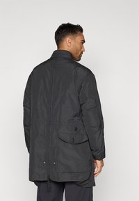 Black nylon jacket with a high collar, buttoned pockets, and a drawstring waistband. Textured fabric with a loose silhouette and button accents.