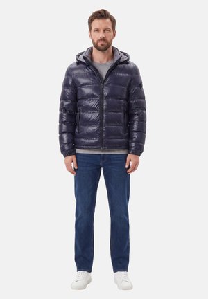 Uomo in piedi che indossa una giacca a vento blu scuro sopra una maglietta grigia, jeans blu e sneakers bianche su uno sfondo chiaro e uniforme.