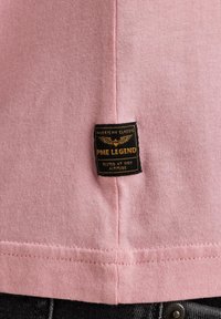 Svijetloružičasta pamučna majica s crnom etiketom na kojoj se nalazi natpis "PME Legend" i dizajn krila. Šavovi su vidljivi duž donjeg rubа.