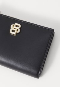 Portafoglio in pelle nera con una texture liscia, caratterizzato da un accento in logo metallico dorato e bordi cuciti. Design rettangolare compatto.