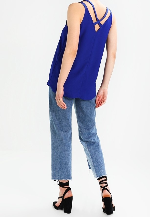 Personne portant un haut bleu sans manches avec des bretelles croisées, un jean court bleu clair et des chaussures noires à talons blocs lacées, debout sur fond blanc.