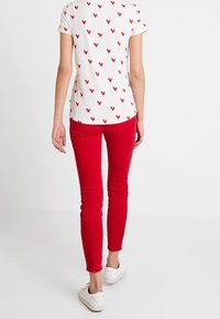 Biała koszulka z czerwonym wzorem serca, zestawiona z czerwonymi, dopasowanymi dżinsami i białymi trampkami. Skupienie na casualowym, lekkim materiale bawełnianym.