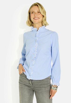 Mujer sonriente con cabello rubio corto que lleva una blusa azul claro con volantes y jeans grises, con pulseras en su muñeca derecha.