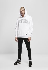 Hvit hettegenser med "NEW YORK" trykt i store svarte bokstaver, frontlomme, kombinert med svarte slim-fit bukser og svarte sneakers.