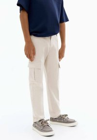 Beige cargobroek met zijzakken, hoge taille en recht pijpdesign. Gecombineerd met grijze schoenen en een donkerblauw shirt met korte mouwen.