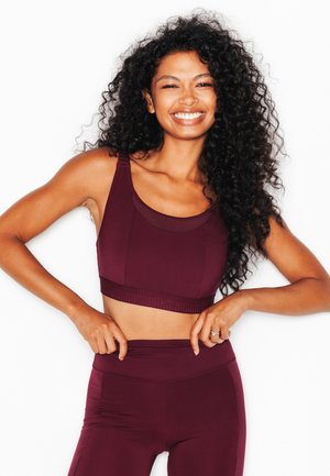 Femme souriante aux longs cheveux bouclés portant un soutien-gorge de sport et des leggings bordeaux assortis, ajustant la taille.