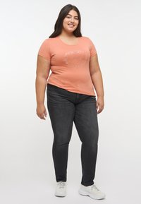 Ljus korallfärgad t-shirt med kort ärm och rund hals, med en silvergrafikdesign; tillsammans med mörka denim slim fit jeans och vita sneakers.