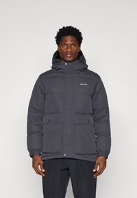 Padded black jacket with a hood, snap closure, two front pockets, elastic cuffs, and a logo on the left chest. Water-resistant fabric.