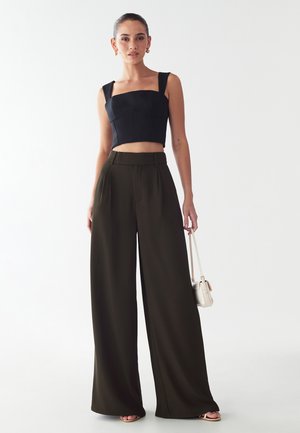 Zwarte crop-top met brede banden, gecombineerd met hoge, wijde olijfgroene broeken. Het model houdt een kleine handtas vast.