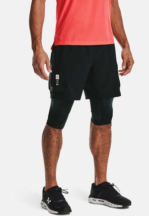 Man die zwarte gelaagde korte broek met zijzak draagt over compressiebroek en zwarte hardloopschoenen, staand tegen een witte achtergrond.